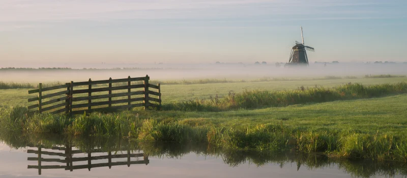 Eine weite Landschaft mit Wasserlauf, Wiese, Nebel und einer Windmühle im Hintergrund. Das Bild steht für die regionale Verwurzelung der Mitglieder des Genossenschaftsverbands Weser-Ems im Norden.