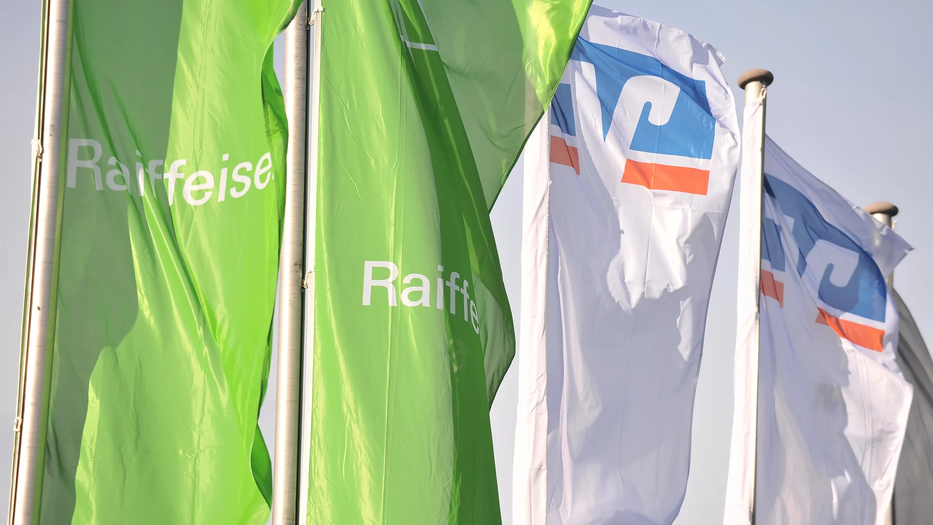 Grüne Raiffeisen-Flaggen und weiße Flaggen mit VR-Logo wehen vor hellem Himmel.